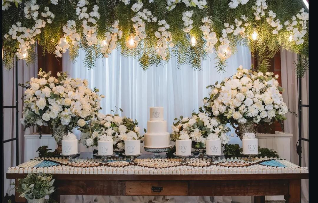 Mesa do bolo com florais brancos suspensos e mini bolos personalizados